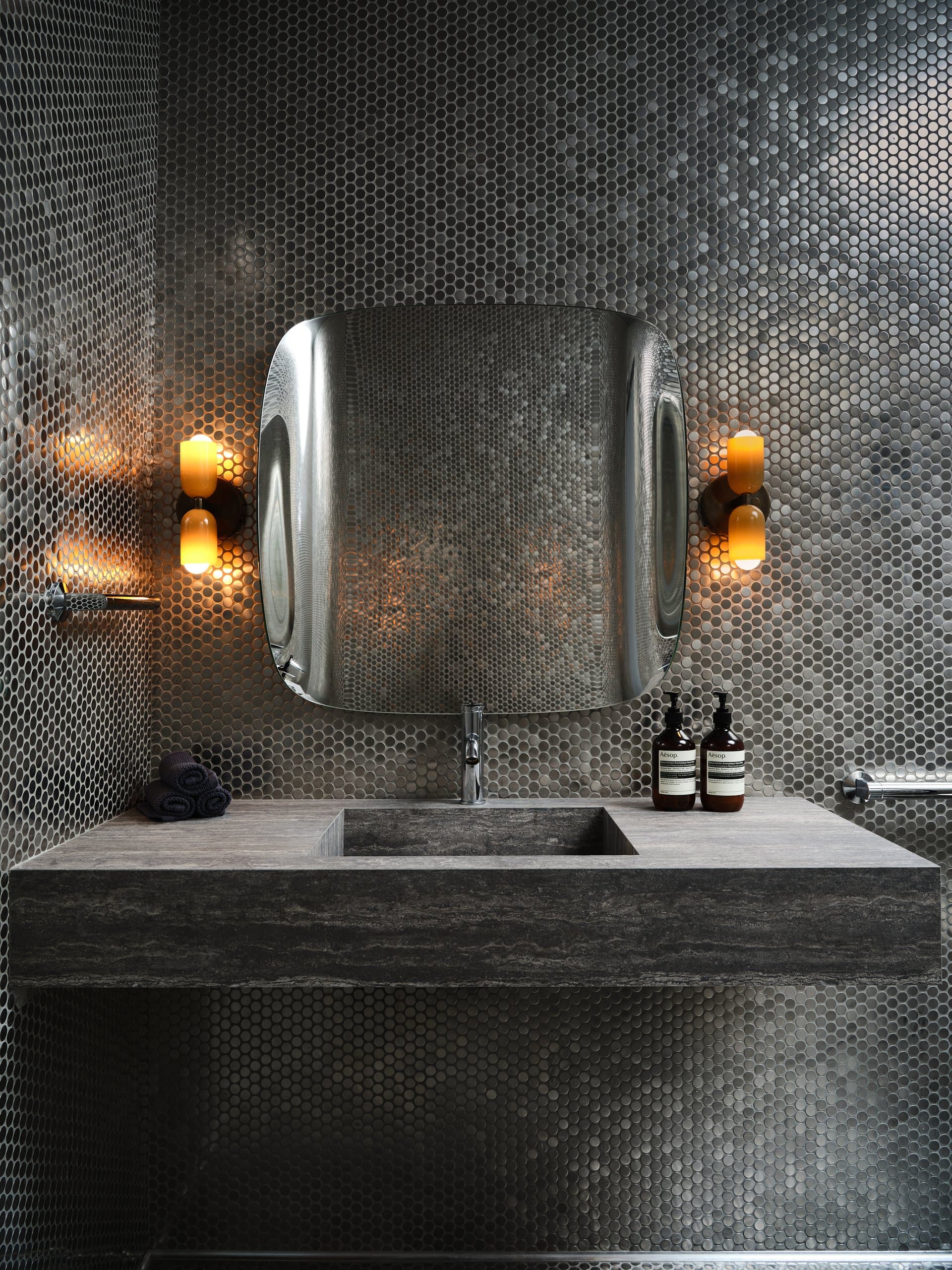 A dark penny-tiled bathroom with an ornate mirror, warm sconces, and a stone vanity